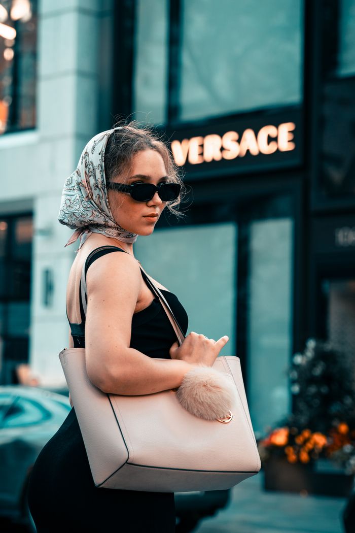 Elegant woman wearing a scarf and sunglasses, posing with a designer bag in a city setting.